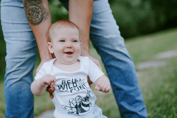Ein lächelndes Baby in einem weißen Strampler mit dem Aufdruck 'Be Wild and Have Fun'. Im Hintergrund steht eine Person, die das Baby von hinten hält, vor einem grünen Rasen. Lächelndes Baby in weißem Strampler mit 'Be Wild and Have Fun' Aufdruck, gehalten von einer Person im Freien.