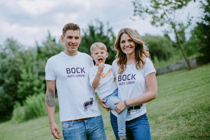 Ein lächelndes Paar mit einem Kind steht auf einer Wiese. Sie tragen weiße T-Shirts mit dem Schriftzug 'Bock aufs Leben'. Familie auf einer Wiese mit 'Bock aufs Leben' T-Shirts